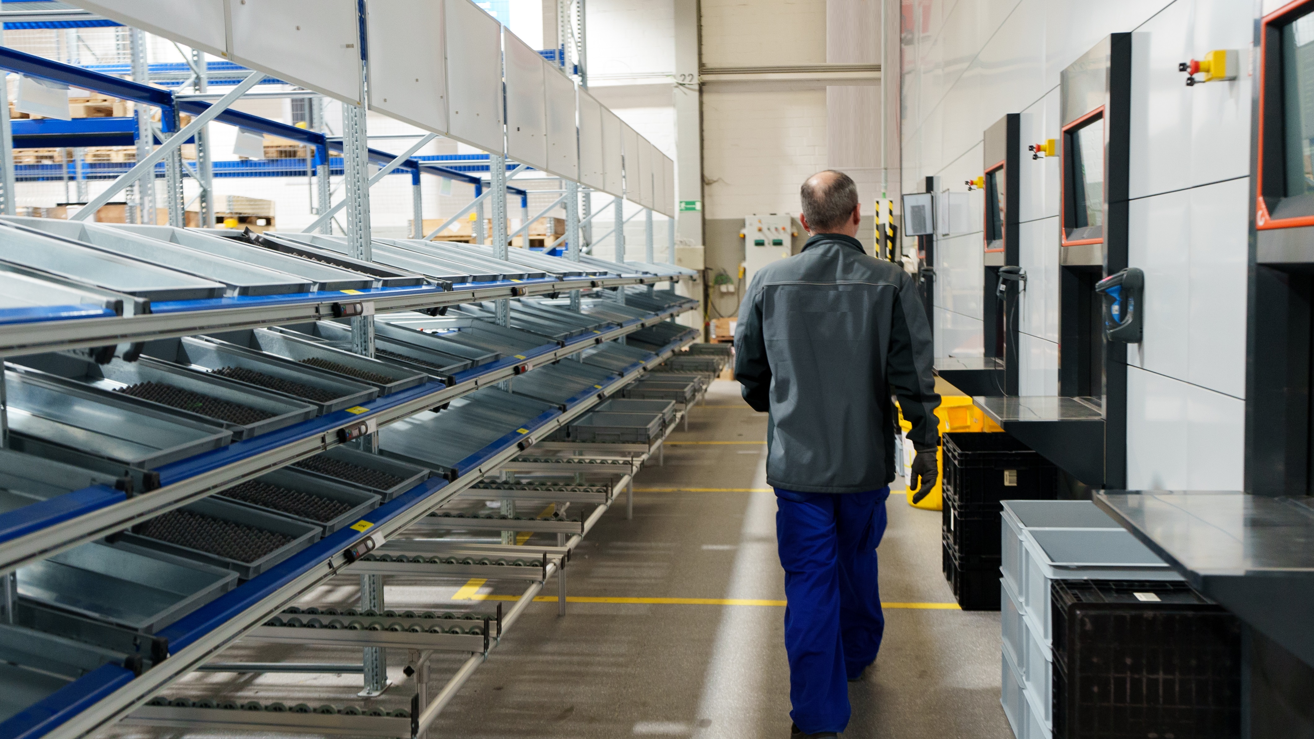 View from behind a worker walking through Demag’s CTO assembly area, showing the Agilon system, pick-to-light stations, and organized flow racks along the production line.